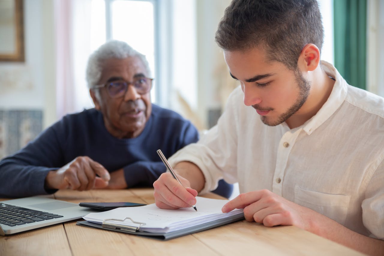 A young man aids a senior citizen with writing at a wooden table indoors, fostering social assistance and connection.